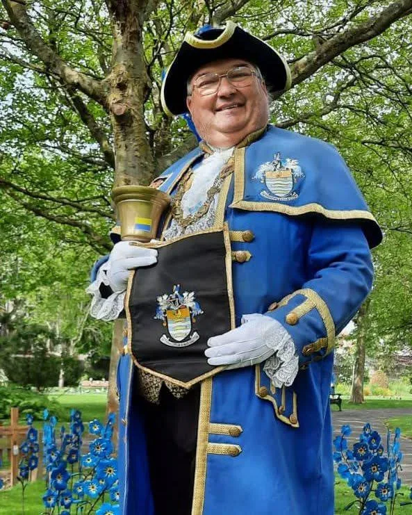 Bob Smytherman, Official Town Crier for Worthing and Adur, in his blue and gold robe and tricorn hat, holding his brass bell in a public garden.